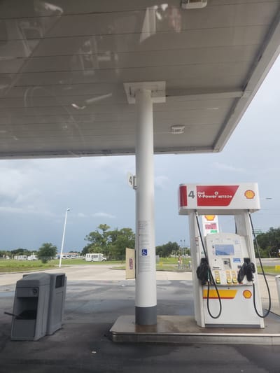 The image depicts a gas station in Merritt Island, United States. A white Shell V-Power Nitro+ fuel pump, labeled with the number 4, is prominently featured on the right side of the frame. The pump has two nozzles and a display screen showing current fuel prices. To the left of the pump, a gray trash receptacle is visible. The scene is under a covered canopy, with a white column supporting the roof structure. In the background, there are trees, utility poles, and several recreational vehicles parked in a lot. The sky is overcast, suggesting potentially stormy weather. A sign with a wheelchair symbol and safety instructions is attached to the white column on the left. The ground appears wet, possibly from recent rain. A street sign indicating "11TH RD" is visible in the far right background.
