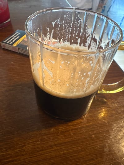 The scene depicts an indoor setting, likely a cafe or bar, located at an unspecified address within the city of Kempten (Allgäu), Germany. The primary subject is a clear, faceted glass mug, approximately one-third full of a dark brown liquid topped with a substantial layer of light brown foam. The foam adheres to the interior walls of the mug, indicating partial consumption of the beverage. The mug rests on a dark brown, polished wooden table exhibiting visible grain. To the left and slightly behind the mug, a white and black cigarette pack is visible, displaying text in Cyrillic script, specifically a health warning in Bulgarian. Further to the left, a portion of a red-colored glass or drink is present. The background shows blurred elements, including what appears to be a person wearing a light blue top. There are no direct interactions captured. The environment is illuminated by ambient light, creating reflections on the glass mug and table surface.