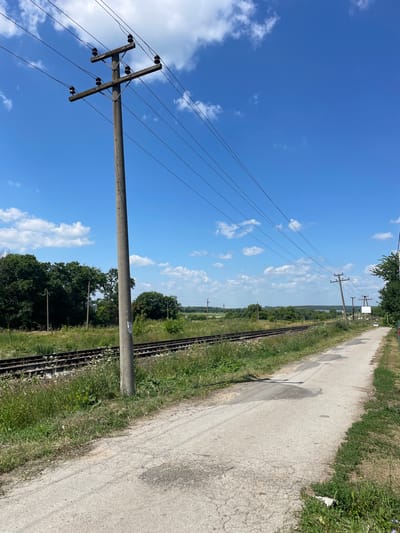 A dirt road runs alongside railroad tracks under a bright blue sky with scattered white clouds. A concrete utility pole with multiple wires stands on the left side of the road. The surrounding landscape is green and rural, with trees and fields visible in the distance. The scene appears to be in the village of Barsuki, Russia.