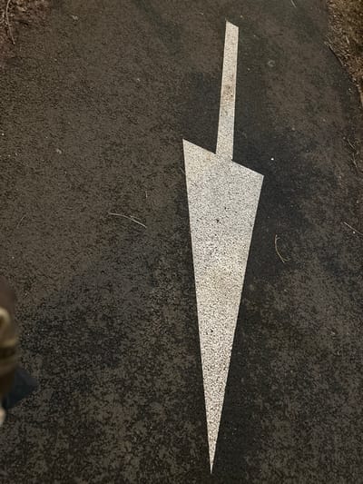 A white arrow is painted on a black asphalt surface. The arrow has a long, narrow head pointing to the right, connected to a shorter, rectangular tail. The paint appears slightly textured and granular. The black asphalt has a rough, worn surface. Stray strands of dried grass or small twigs are scattered around the arrow. Part of a person's leg and shoe are visible in the bottom right corner.