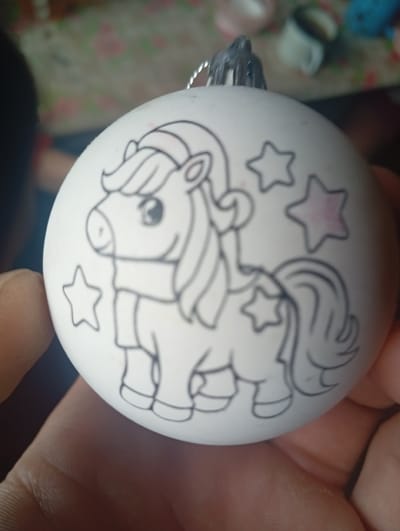 A white spherical Christmas ornament featuring a black outline drawing of a unicorn with stars is held in a person's hand. The unicorn is depicted with a mane, tail, and wearing a scarf. Some of the stars have a subtle pink hue. The ornament has a silver hanging loop. The background is out of focus and appears to be an indoor setting with a floral tablecloth and various objects. The image does not contain any information related to the city of Bryansk, Russia.