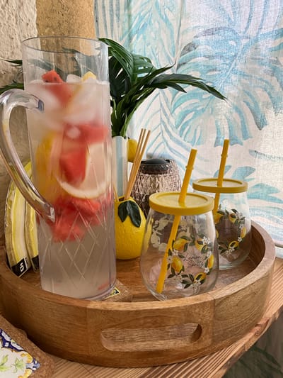 The image shows a close-up of a wooden tray holding a pitcher of lemonade with fruit and ice, two glasses with yellow lids and straws, a decorative lemon-shaped object, and some napkins. The pitcher is clear plastic with a diamond pattern and contains lemonade with slices of watermelon and lemon, as well as ice cubes. The two glasses are decorated with lemons and the words "Lemon Spritz" and have yellow lids with yellow straws inserted. To the left of the pitcher are yellow and white striped napkins. To the right of the pitcher is a decorative yellow object shaped like a lemon with green leaves, and behind it is a vase with wooden stir sticks and a decorative textured object. A green palm frond is visible behind the pitcher. The background features a light-colored curtain with a blue tropical leaf pattern. The scene appears to be set indoors, possibly on a table, given the wooden surface beneath the tray. There is no visible indication of the location in Memmingen, Germany.