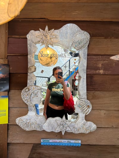 The image shows a woman taking a selfie in a mirror in Suthep, Thailand. The woman is wearing a green crop top and black pants. She is holding a red bag in her left hand. The mirror is decorated with a star and clouds. Behind the mirror, there is a wooden wall and a staircase. On the left side of the mirror, there are two books. The overall scene suggests a casual, possibly artistic or bohemian setting, typical of some cafes or boutiques found in tourist areas of Suthep.