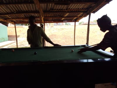 Two individuals, silhouetted against the bright daylight filtering through a corrugated metal roof supported by wooden posts, are engaged in a game of billiards. The scene is set outdoors, under a makeshift shelter, on what appears to be a dirt ground. In the foreground, a green billiard table is positioned. On the table, there are three visible balls: a dark-colored ball, an orange-colored ball, and a white cue ball. One individual, positioned on the left, is standing and appears to be observing the game. The other individual, on the right, is actively playing, holding a cue stick and aiming at the balls. The background reveals a rural landscape with some buildings and vegetation, suggesting a location outside of a densely populated urban area. The overall ambiance is casual and recreational. The location is identified as Kachia, Nigeria.