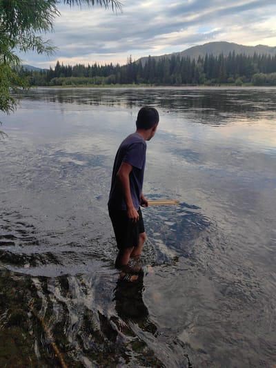 A young male wearing a dark t-shirt and black shorts stands in the shallow water of a wide river. He is holding a long, light-colored object, possibly a stick or fishing pole, which extends into the water. The river is calm with ripples reflecting the cloudy sky and the forested shoreline in the background. The trees are dense and dark green, and a distant mountain range is visible under a pale sky with some soft clouds. The scene appears to be in a natural, rural setting.