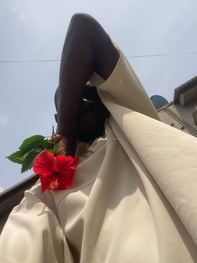 The image displays an individual outdoors in Langbasa, Nigeria, captured from a low-angle perspective looking upwards towards a bright sky. A dark-skinned person, wearing a light beige collared shirt, occupies the foreground. Their right forearm is raised, obscuring the upper portion of their face, while the lower face, including an open mouth with visible teeth, is discernible in shadow. A vibrant red hibiscus flower, complete with green leaves, is held by the person's left hand and positioned against their chest. The background features a clear, light blue sky with a faint horizontal wire visible. On the upper right, sections of a building are present, including a wall and a grey satellite dish. The scene is illuminated by strong sunlight, casting distinct shadows.