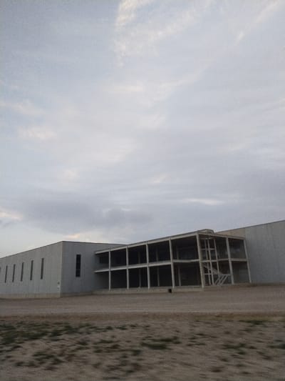A low-angle shot captures a large, rectangular building under a cloudy sky. The building features a long, windowed side section leading to a two-story, open-frame structure. Adjacent is a solid, windowless section. The terrain is sandy with sparse vegetation. The scene suggests a construction site or industrial area with overcast lighting.
