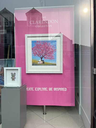 The image depicts a storefront window display at 11 High Street, Esher, in the Borough of Elmbridge, United Kingdom, specifically a Clarendon Fine Art gallery. The interior display features a large, bright pink banner serving as a backdrop. "THE CLARENDON COLLECTION" is printed in white capital letters at the top of the banner, and "CREATE. EXPLORE. BE INSPIRED" is printed in white capital letters at the bottom. The banner is supported by two visible chrome posts at its base.
Two artworks are displayed. A larger, square-format painting in a white wooden frame is centrally positioned against the pink backdrop. The painting depicts a vibrantly pink-blossomed tree in a field of yellow flowers under a bright blue sky. Two stylized figures, one light-colored and one darker, are seated side-by-side beneath the tree. The artist's signature, "Gary Walton," is visible at the bottom center of the painting.
To the left of the main painting, a smaller, white-framed, square-format artwork rests on a dark grey rectangular pedestal. This piece contains a three-dimensional, multi-colored mosaic-style teddy bear figure on a white background.
The reflective glass window captures elements of the external environment. A prominent reflection in the upper left shows a multi-story building with light-colored masonry and an ornate cupola or turret. Other reflections include reversed text from adjacent businesses, such as "TREASURES" and "ONE SERVICE CENTRE," visible on the right side of the glass. A bright sun glare is present in the upper right corner of the window. The interior floor visible is composed of light-colored square tiles.