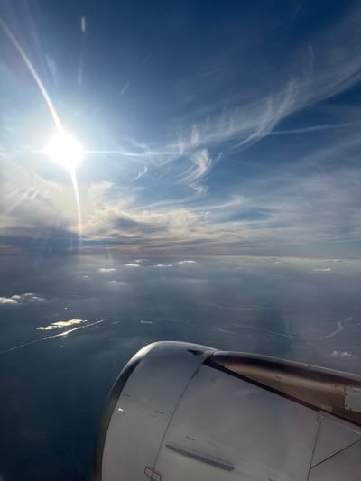 This image presents an aerial view from an airplane window, showing a bright sky, distant land, and part of the aircraft's engine and wing. The location context indicates the flight is above or near Combs-la-Ville, France.
The upper and left portions of the frame are dominated by a clear, deep blue sky, accented by delicate, wispy white clouds that stretch horizontally across the expanse, resembling cirrus clouds or high-altitude contrails. In the lower-left, a brilliant sun radiates intense light, causing significant lens flare and bright sunbeams that extend across the sky, highlighting the cloud structures.
A discernible horizon line runs across the mid-right section of the image, separating the sky from the terrestrial landscape below. The ground appears as a dark, somewhat hazy landmass, with scattered lighter patches that likely represent reflections from water bodies or distinct terrain features, illuminated by the sun.
Prominently featured in the upper-right corner is a portion of the airplane's engine nacelle and wing. The engine is a large, metallic structure, primarily white with darker sections, and exhibits visible rivets and panel lines. Small, dark text markings are visible on its surface, including "HOT POINT" repeated in several places, and a small red rectangular marking with unreadable white text, possibly a warning or instruction label.
The scene suggests a bright daylight setting, likely late morning or afternoon, given the sun's strong glare. The weather appears clear with some high-level cloud formations.