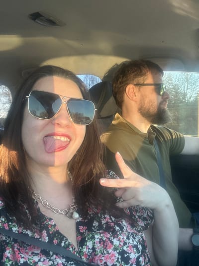 Two people are in a car. A woman in the foreground is wearing large sunglasses, a floral shirt, and a silver chain necklace. She is sticking her tongue out. A man behind her is wearing sunglasses and a green shirt. A hand is raised in a peace sign. The location is unknown.