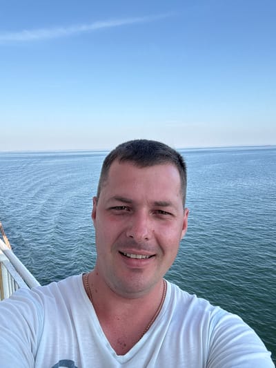 A man with short, dark hair and brown eyes smiles at the camera. He is wearing a white t-shirt and a thin gold chain. The photo is a selfie, taken from the chest up, with the man's face turned slightly towards the camera. He is outdoors, with a bright blue sky and a body of water behind him. The water appears calm with slight ripples. The sun is shining, suggesting it is daytime, likely late afternoon or early evening given the soft light. The man's expression is relaxed and happy. There are no visible objects other than the man and his clothing. No text is visible in the image.