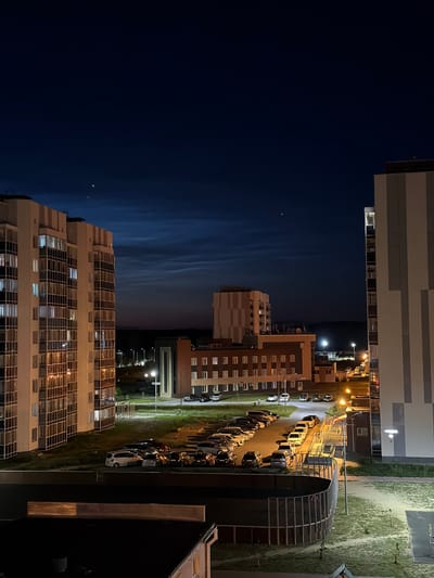 The image displays a nighttime scene in Tsiolkovsky, Russia, featuring apartment buildings, a parking lot filled with cars, and streetlights. The perspective is from an elevated vantage point, looking down and across the scene. The sky is dark blue with faint, wispy clouds, suggesting it is either dusk or pre-dawn. A few distant lights are visible in the sky, possibly stars or aircraft.

The buildings are modern, multi-story structures with many windows, some of which are illuminated, indicating occupancy. One building in the foreground has a facade with a grid-like pattern of balconies or windows. Another building, further in the distance, has a more segmented facade with contrasting color blocks.

A row of parked cars lines a paved area next to a grassy strip, illuminated by bright streetlights. A distinctive feature is a curved, glass-enclosed structure, possibly a bus stop or a modern architectural element, with visible internal lighting and supports. The overall atmosphere is quiet and residential, characteristic of a suburban or urban nighttime setting. No people are explicitly visible, and no text is discernible in the image. The lighting suggests artificial illumination from street lamps and building interiors, casting shadows and highlighting the architectural features.