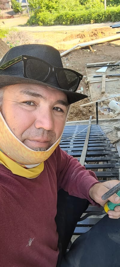 The image shows a close-up, rotated selfie of a man, likely in Milisi, Georgia. The man is wearing a burgundy sweatshirt, a black hat with sunglasses perched on top, and a yellow patterned cloth around his neck, possibly a mask. His expression is neutral, and his gaze is directed towards the camera. In his right hand, he holds a tool with a green and yellow handle, suggesting he might be engaged in some form of construction or repair work.
The background reveals a construction site with scattered building materials, including metal bars, wooden planks, and dirt. A fence made of vertical metal bars is visible behind him, and a green, leafy area with some trees and vegetation is present to the left. The lighting suggests it is daytime, likely sunny. The overall scene implies an outdoor work environment. There is no visible text in the image.