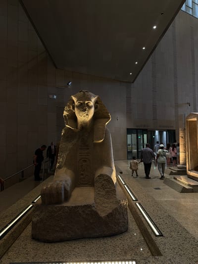 The image shows a large stone sphinx statue in what appears to be a museum or archaeological site. The sphinx is illuminated by spotlights from above, casting dramatic shadows. In the background, several people are walking, including adults and children. There is a set of stairs with a railing to the left of the sphinx, and a doorway with signage that reads "ARCHAEOLOGICAL STUDY" in English and Arabic. The environment appears to be indoors, with tiled walls and a high, angled ceiling. The floor is a speckled granite. Based on the context of the sphinx statue and the "ARCHAEOLOGICAL STUDY" signage, the location is likely within the Grand Egyptian Museum in Giza, Egypt, as Kafrat Nassar is a district within Giza.