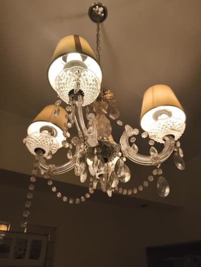 The image shows a chandelier with three lampshades and crystal accents. The lampshades are beige with a dark band at the top. The light is warm, suggesting a residential setting. The location is not discernible from the image.