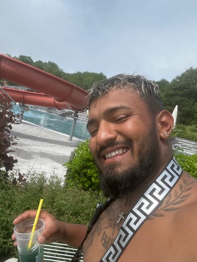 A man is pictured outdoors, likely at a recreational water facility in Oberstenfeld, Germany. He is holding a clear plastic cup with a green beverage and a yellow straw. He has a beard and lightened hair. He is wearing a shoulder strap with a Greek key design and a necklace with a cross pendant. A waterslide is visible in the background next to a swimming pool and greenery. The sky appears overcast.