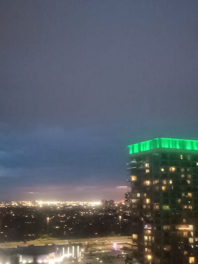 An exterior, medium-shot, eye-level, wide-angle photograph captures a nighttime cityscape. The sky is dark and cloudy, with a band of lighter gray and blue clouds stretching across the middle of the frame. A tall, modern skyscraper with a luminous green rooftop is positioned on the right side of the image. The building has numerous illuminated windows, suggesting occupancy. In the background, a vast expanse of city lights twinkles, indicating a densely populated urban area. The lights create a hazy glow in the distance, typical of a metropolitan setting at night. The foreground shows a lower-lying structure with bright, horizontal lights, possibly a roadway or another building. The overall scene evokes a sense of an active city after dark, with the glowing skyscraper serving as a prominent focal point.