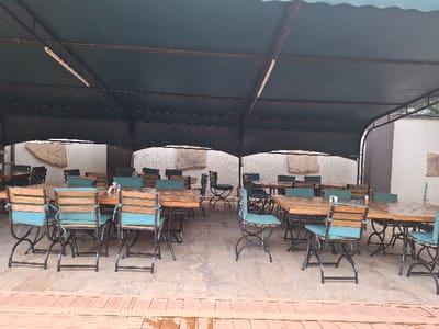 The image shows an outdoor dining area in Karagita, Kenya, located at coordinates -0.754 latitude and 36.438 longitude.  The area is covered by a dark green awning supported by metal poles. Multiple tables and chairs are arranged in rows. The tables are made of wood and have blue cushions on the chairs. The floor is made of light-colored concrete and there is a wooden platform in the foreground. There are no people present in the scene.  The environment is clean and well-maintained.  The chairs are empty and the tables are clear, suggesting that the area is closed or awaiting customers.  The awning provides shade from the sun.  The scene appears to be a casual dining area for a restaurant or hotel.