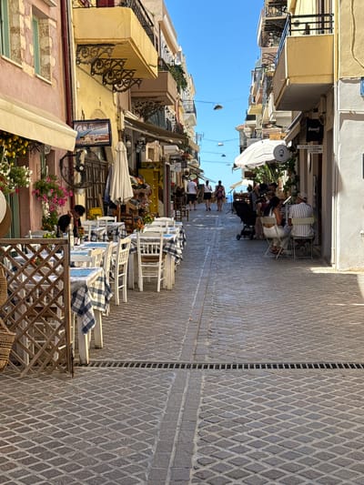 The image captures a narrow, cobblestone street in Chania, Greece, lined with buildings and outdoor dining establishments. On the left, a pink building with arched windows and a cream-colored awning is visible. A sign for "Okeanida Greek Restaurant" hangs on a yellow building further down the street. Several tables with blue and white checkered tablecloths are set up on the right side of the street, occupied by diners. A person wearing a black shirt sits alone at one of the tables. Further down the street, more tables and people are seated, some under a large white umbrella. The street is paved with diamond-shaped cobblestones, and a metal grate runs across the middle. The sky is a clear, bright blue. The overall scene depicts a vibrant, Mediterranean atmosphere, typical of the old town in Chania.