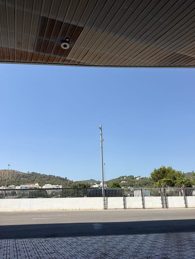 This media file, taken in Sant Antoni de Portmany, Spain, depicts an outdoor scene under a clear blue sky, suggesting daytime with good weather. The image is vertically oriented and appears to be shot from a low angle, looking upwards.

On the left side of the frame, a large, corrugated metal wall dominates, with visible rust streaks and a circular object attached to it, possibly a light fixture or security camera. This wall suggests an industrial or utilitarian structure.

The center of the image is filled with a vast expanse of bright blue sky. A solitary lamppost with multiple fixtures stands vertically in the sky, breaking the uniformity of the blue.

To the right, a paved road runs parallel to a raised concrete barrier topped with a metal railing. Beyond the railing, a densely vegetated hillside is visible, dotted with buildings that suggest a town or village nestled into the landscape. The hillside is covered in trees and shrubbery, and some white buildings with dark roofs are discernible. A patterned tiled sidewalk runs along the very edge of the road on the right.

The composition creates a strong division between the man-made structures of the wall and road, and the natural hillside and sky. There are no people visible, and no specific activity is taking place. The overall impression is one of an urban or semi-urban environment adjacent to a natural, hilly landscape, captured on a bright, clear day.