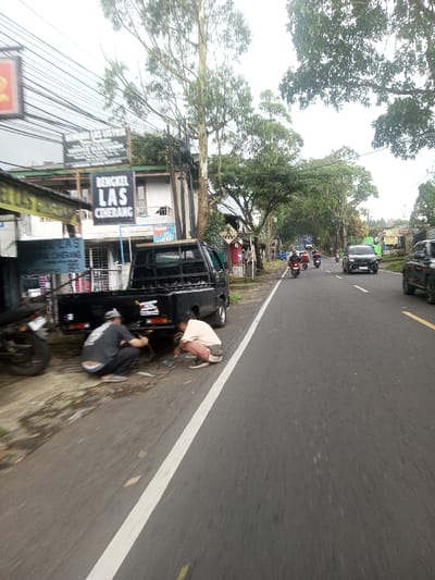 On a roadside, two men work on a black truck, their figures crouched near the rear. A motorcycle is parked behind the truck. The scene unfolds along a road marked with a white line, leading to distant traffic including cars and motorcycles. Buildings and trees border the road, contributing to a busy street scene under an overcast sky. Signage and overhead wires are visible.