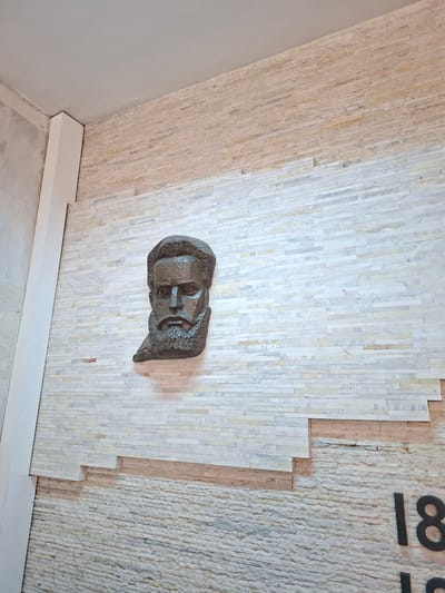 The image shows a bronze bust of a man mounted on a wall covered in vertically-oriented, narrow tiles of varying off-white shades. The wall appears to be part of an indoor space, as indicated by the presence of a white baseboard along the bottom. To the right of the tiled section is a different type of wall covering, possibly plaster or another type of tile, with the partial number "18" visible. The location is the House of Culture "Probuida-1927" in Nova Zagora, Bulgaria, specifically on the wall of the cafe.