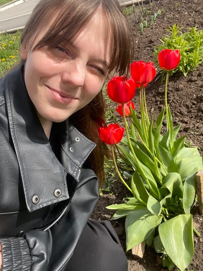 A young woman is lying down in a garden, next to a bunch of red tulips. She is wearing a black leather jacket and black pants. She is smiling and has her eyes closed. The background includes soil, grass, and other plants. The scene is outdoors. The location is not evident in the image. The city of Urussu, Russia, cannot be confirmed from this image.