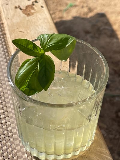 A clear, fluted glass containing a light yellow-green iced beverage is depicted, garnished with a sprig of fresh basil leaves. The glass rests on a light-colored woven surface, with a light wooden plank visible behind and to its right. The background consists of blurred, reddish-brown ground, consistent with an arid environment. The scene is brightly illuminated by direct sunlight, casting distinct shadows. This location is the Arava Tichona Regional Council, Israel. No individuals or active interactions are present.