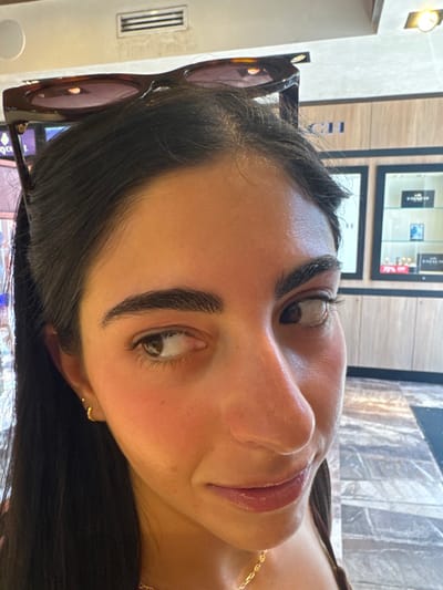 The image presents a close-up, head-and-shoulders view of an individual, likely female, with dark hair and a light complexion. Dark brown tortoiseshell sunglasses rest on top of the subject's head. The subject's face is oriented slightly towards the right, with both brown eyes directed towards the viewer's left. The mouth is slightly agape, revealing glossy lips. A small gold hoop earring is visible on the left earlobe, and a thin gold chain necklace is worn around the neck. The background reveals an indoor commercial environment. A rectangular ventilation grate is visible on the upper left wall or ceiling. To the right, the word 'COACH' is displayed on a light-colored wall panel, indicating a retail store. Below this, two framed display cases contain commercial products, one of which includes a partial '70% OFF' price reduction sign. The floor in the lower right consists of light-colored, patterned tiles. This scene is consistent with a retail interior, such as those found in the commercial districts of San Miguel de Cozumel, Mexico.