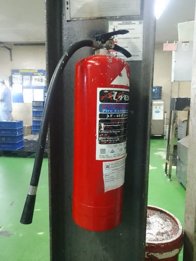 A red fire extinguisher is mounted on a black pillar in what appears to be an industrial or warehouse setting. The fire extinguisher has a black hose and handle. There is a label on the extinguisher with text and graphics, including the brand name "Aire." Above the fire extinguisher, a white sign with text and graphics is attached to the pillar, which appears to be a set of instructions for using the fire extinguisher. In the background, there are blue crates stacked on a pallet, a water cooler, and a person wearing a light-colored uniform. The floor is green. The overall environment suggests a functional space, possibly a factory or storage area, located in Ciantra, Indonesia.