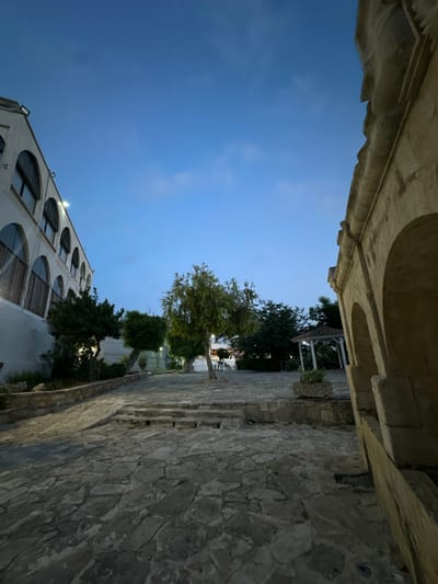 The image depicts a courtyard area in Peyia, Cyprus, during twilight. A white building with arched windows lines the left side of the scene, while a stone structure with arches dominates the right foreground. The ground is paved with irregular flagstones, and a set of stone steps leads up to a slightly elevated area in the center. Several trees with lush green foliage are scattered throughout the courtyard, with a small white gazebo visible in the background. The sky is a clear, deep blue, transitioning to a lighter hue near the horizon. No people are visible in the image. The lighting suggests it is either dusk or dawn, with artificial lights illuminating parts of the building and the courtyard.