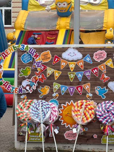 Here is a description of the image:

The image shows a children's party or event. A large, colorful inflatable bouncy castle with a Minion theme is visible on the left. In front of the bouncy castle is a wooden wall decorated with various cartoonish decorations, including cloud shapes, butterflies, birds, bees, and a cartoon child on a skateboard. There are also triangle-shaped flags with letters on them, forming part of a celebratory message. Large, wrapped lollipops on sticks are attached to the wall. Banners or signs with Russian text are scattered around the wall, some emphasizing "Children's Day". The environment appears to be outdoors, possibly at a park or event space. The location, Tokkari-Lend, Russia, is not apparent from the image itself but might be inferred based on the content or other clues.