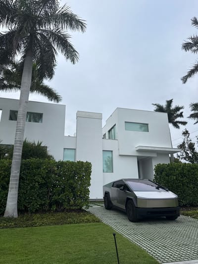 The image displays an exterior scene featuring a modern residential property in Naples, Florida, United States of America. A grey and silver Tesla Cybertruck is parked diagonally on a light-colored grid paver driveway, positioned in front of a multi-level, contemporary white stucco house. The residence exhibits sharp angles, flat roofs, and numerous rectangular windows, some appearing tinted or reflective. Green, manicured hedges line the perimeter of the property and the base of the house, separating it from a vibrant green lawn in the foreground. Several tall palm trees with grey trunks and green fronds are prominent features, framing the house against an overcast, light grey sky. No individuals are present in the scene.