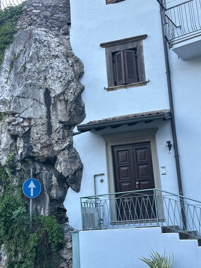 The image captures a narrow street in Cividate Camuno, Italy, dominated by a dramatic rock face that juts out precariously close to a building. The building, painted a pale blue, features a dark wooden double door with a traditional Italian design, accessed by a few steps and a mint green metal railing. Above the door, a small, dark fixture hangs on the wall, possibly a light or decorative element. A window with brown shutters is visible higher up on the building. Green ivy climbs the rock face and the side of the building, softening the stark contrast between nature and architecture. A blue circular traffic sign with a white arrow pointing upwards is affixed to a pole on the left side of the image, indicating a one-way street. The overall scene suggests a town built into its natural landscape, with elements of both ruggedness and residential charm.