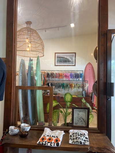 The image shows the interior of a surf shop, viewed through a glass door. The shop is located at the coordinates 9.644 latitude and -85.167 longitude, in the city of Santa Fe, Costa Rica. The shop is brightly lit, with a large wicker lamp hanging from the ceiling. The walls are painted white, and the floor is made of wood. There are several surfboards leaning against the wall, including one with the word "Holal" written on it. A rack of colorful clothes hangs behind the surfboards. To the right of the surfboards, a large plant in a wooden pot stands in the corner of the room. In the background, a framed photo of a wave hangs on the wall. A photo frame sits on a wooden table in the center of the shop, featuring a portrait of a woman with long, dark hair. Two white t-shirts with the phrase "Calma, Respeto, Amor" printed on them lay on the table. The t-shirts are on a wooden surface, positioned on a small table outside the shop. The glass door is open, and a portion of a dark shirt is visible in the bottom left corner of the image.