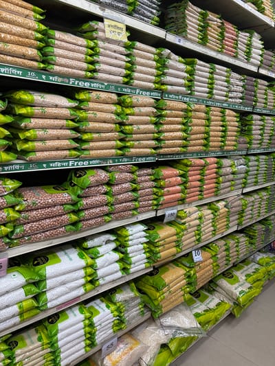 The image displays a detailed view inside a retail grocery store located in Ambernath, India. The scene is dominated by multiple levels of white metal shelving units, fully stocked with various packaged dry food items. Numerous clear plastic bags, predominantly featuring green and white branding (partially identifiable as "Indian Choice"), contain different types of legumes, pulses, and grains, including yellow split peas, white tapioca pearls (sabudana), whole peanuts, and red lentils. These packages are uniformly stacked horizontally and vertically across at least five visible shelves. White price tags with numerical values such as "150," "32," "55," and "10" are affixed to the front edges of the shelves. Green promotional strips with Marathi text, notably "शॉपिंग 10% SAVINGS १ महिना Free" (Shopping 10% Savings 1 Month Free), are present along the shelves. A yellow sign displaying "अनपॉलिश्ड तुअर दाल" (Unpolished Toor Dal) in Marathi is also visible on an upper shelf. The environment is well-lit, suggesting standard overhead retail lighting. No individuals are present in the frame.