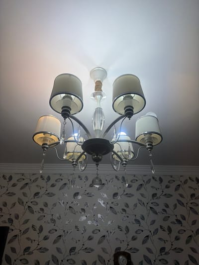 A chandelier hangs from a white ceiling, its fabric shades casting a warm glow. The chandelier is made of dark metal with crystal accents. The wallpaper on the right side of the image features a delicate leaf pattern in shades of grey and white. A small clock with a wooden frame is visible on the wallpaper. The scene appears to be indoors, possibly in a dining room or living room. The lighting suggests it could be evening or a dimly lit day. No people are visible in the image.