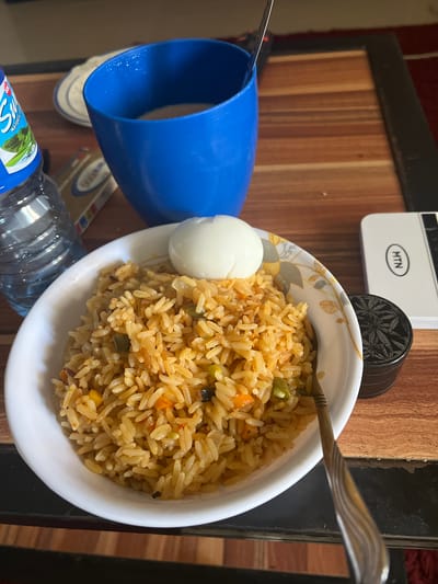 The image shows a close-up of a bowl of fried rice with a hard-boiled egg, placed on a wooden surface. A blue plastic cup and a bottle of water are also visible in the foreground. The fried rice appears to be a mixture of rice, vegetables like carrots and green beans, and possibly other ingredients that give it a yellowish-brown color. A fork is partially submerged in the rice, suggesting it's ready to be eaten.
In the background, a white MTN device is visible, which is a telecommunications company prevalent in Nigeria. This, combined with the "Laminga, Nigeria" location context, suggests the scene is set in Nigeria. There is no indication of time of day or weather. The overall mood is casual and domestic, implying a mealtime. The focus is on the food, presented in a simple, unpretentious manner. The lighting suggests it might be indoors, possibly in a kitchen or dining area. The wooden surface has a distinct grain pattern, adding a rustic touch to the composition.