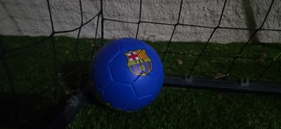 This media file shows a blue soccer ball with the FC Barcelona logo on it, resting on a patch of grass next to a soccer net. The ball is positioned slightly off-center, with the net running across the top portion of the image. The grass appears to be slightly damp, suggesting recent rain or dew, and some fallen leaves are scattered on it. Behind the net, a textured, light-colored wall is visible, likely part of a building or boundary.
The lighting in the image suggests it was taken at dusk or dawn, or possibly under artificial light, as the scene is dimly lit but not completely dark. The overall mood is quiet and still, with no people present. The setting appears to be an outdoor sports area, possibly a backyard or a small field. The presence of the net and ball clearly indicates a context related to soccer. No specific activity is taking place, as the ball is stationary. The visible text on the ball is "FCB", part of the FC Barcelona crest. There are no other discernible text elements. The location context provided is Hadera, Israel.