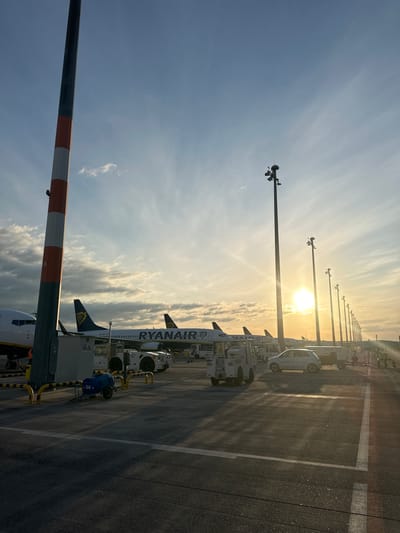 At sunset, a tarmac at Berlin Brandenburg Airport (BER), located in Schönefeld, Germany, is depicted. Several Ryanair airplanes are parked at gates. The sun is low in the sky, casting long shadows and creating lens flare. Tall light poles with multiple lamps line the tarmac, and various airport vehicles, including a small white car, baggage tugs, and a blue water cart, are visible. A large striped pole with a number "6" on it is in the foreground on the left. The sky is partly cloudy with a gradient of blue and orange hues.
