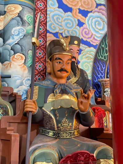 A low-angle, close-up shot depicts a colorful, richly decorated statue of a Chinese warrior figure. The statue is positioned vertically. The warrior has tan skin, a black mustache, and a serious expression. He wears elaborate gold and black armor, and a headdress with a gold animal emblem. The statue is holding a long, dark pole weapon horizontally in both hands.

Behind the warrior, there is a background of painted clouds and other decorative elements in blues, whites, and golds. Another figure is partially visible behind and to the side of the main warrior statue. The second figure has a black hat and a similar tan skin tone.

The statue appears to be located in the city of Lintong District, China.
