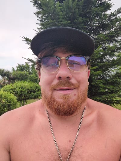A selfie of a man is taken outdoors, with lush green trees and a cloudy sky in the background. The man, who has a reddish-brown beard and is wearing a black baseball cap and sunglasses, is looking directly at the camera. He is shirtless and has a silver chain around his neck. The photo is taken at a close-up range, focusing on the man's face and upper body. The setting appears to be a residential area, possibly a backyard, in the village of Perliavka, Ukraine, given the context.