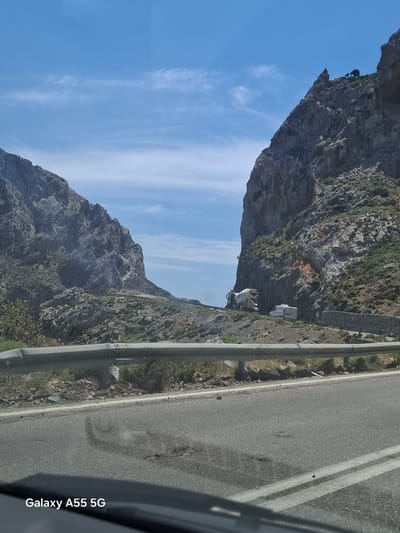 The image, taken on a Galaxy A55 5G, shows a mountain road near Frati, Greece. The road runs along the side of a steep, rocky mountain with sparse vegetation. A gray metal guardrail separates the road from the mountain's edge. Two vehicles, possibly recreational vehicles, are parked or traveling on the road further up the mountain. The sky is blue with wispy white clouds. The foreground shows a portion of the road and possibly part of the vehicle from which the photo was taken.