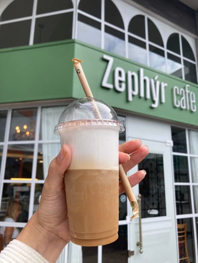 A hand holds a plastic cup of iced coffee in front of a building. The coffee cup is clear with a domed lid. A tan paper straw sticks out of the cup. The drink appears to be layered, with a creamy white layer on top and a tan coffee layer below. The hand holding the cup is pale with neutral nail polish and wearing a white textured sleeve.
Behind the hand and coffee, the green facade of a building is visible. The sign reads "Zephyr cafe" in silver letters. The building has white-framed windows with arched tops, and a white door with a window can be seen.
The location is Florianópolis, Brazil, as evidenced by the "Zephyr cafe" sign, a known business in the area.