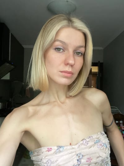 The image shows a young woman with blonde hair and light skin, wearing a pink floral strapless top. She is looking at the camera with a neutral expression. The image was taken indoors in a kitchen or dining area. The woman is standing in front of a wall and her right arm is outstretched. Her left arm is by her side. The background includes an overhead light fixture, a black kitchen cabinet, and a door that is partly open. The image appears to be a selfie, taken by the woman holding a phone or camera. There are no other people in the image. The coordinates provided indicate the location is in the city of Moscow, Russia.