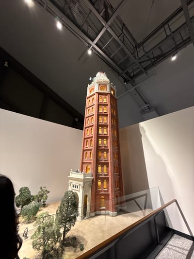 The image depicts a scale model of a tall, red-brick building with numerous windows and white decorative elements at its base and top, possibly inspired by the Tokyo Skytree, although I'm unsure of the exact building this is. The model is enclosed in a glass case. In the background there's a ceiling-mounted industrial metal structure with lights. The diorama base of the model features miniature trees and figures. A small portion of a person's head is visible at the bottom of the frame.