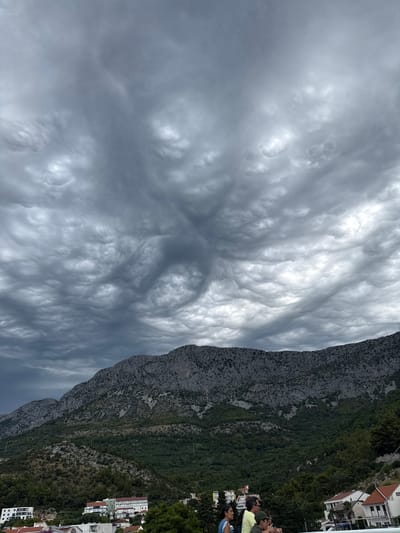 This is a landscape photograph taken from Drvenik, Croatia, showcasing a dramatic sky filled with mammatus clouds over a rugged mountain range. The clouds are dense and gray, with a distinctive undulating pattern, suggesting an impending storm. Below the imposing sky, the steep, rocky slopes of the mountain are covered in patches of green vegetation. Nestled at the base of the mountain are several buildings, indicating a residential or tourist area. In the foreground, partially visible, are a group of people, seemingly observing the sky. Their presence suggests they are either residents or visitors enjoying the view of the unusual cloud formation. The overall atmosphere is one of awe and a touch of foreboding due to the intense cloud display.