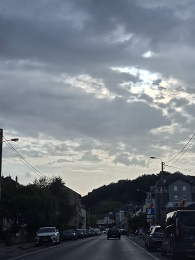 The image shows a street scene in Sanem, Luxembourg, under a cloudy sky. A row of parked cars lines the right side of the road, with a dark-colored van prominently in the foreground. To the right, a Q8 gas station is visible with illuminated price boards displaying fuel costs. Buildings, including residential structures and what appears to be a gas station convenience store, are situated behind the gas pumps. Trees and foliage are present along the left side of the street, with more buildings and a hillside covered in dark trees forming the background. The road itself is paved and appears to be a main thoroughfare. The lighting suggests it is either early morning or late evening, with limited sunlight breaking through the clouds.