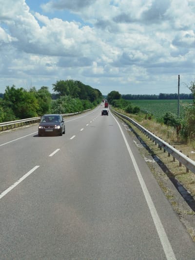 The image shows a view from a car driving on a highway. The highway has two lanes in each direction, separated by a median. There are guardrails on the side of the road, and trees and fields line the highway. In the foreground, there is a dark-colored car driving in the left lane. In the distance, there are other cars and trucks on the highway. The sky is blue with some clouds. The location appears to be a rural area, possibly on the outskirts of a city. The image is taken from a slightly elevated position, looking down the road. The license plate of the car in the foreground is partially visible. There are no people clearly visible in the image, besides the driver in the foreground car. The scene appears to be peaceful and ordinary, with normal traffic flow. The date is not provided, but the weather suggests a warm day.