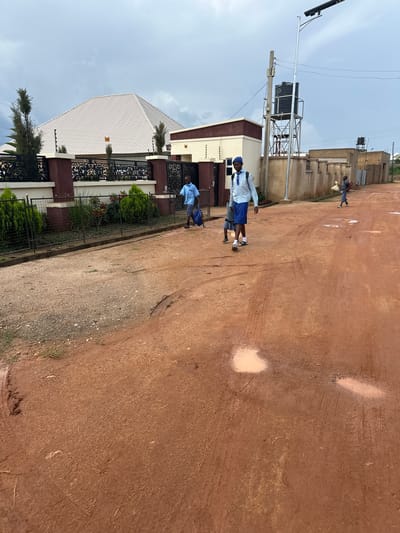 This image depicts a street scene in Rayfield, Nigeria, with a focus on two young people, likely students, walking on a dirt road. The road surface is reddish-brown and appears to be a mix of dirt and gravel, with visible tire tracks and puddles, suggesting recent rain or a naturally wet area. The sky is overcast, indicating a cloudy day.

One individual, wearing a light blue shirt, dark shorts, and a blue cap, is walking towards the viewer. They are carrying a blue bag over their shoulder and appear to be wearing sneakers. Another person, further up the road and partially obscured, is also walking away from the viewer, carrying a lighter colored bag and wearing a blue and white outfit.

In the background, to the left, is a residential building with a white, corrugated metal roof and decorative black metal fencing. Adjacent to this is another building with a light yellow facade. A large water tank on a metal stand is also visible, suggesting a potential water supply system. The overall setting is a semi-urban or rural environment in Nigeria, characterized by the unpaved road and the style of the buildings. The time of day is indeterminate due to the overcast sky, but the scene suggests an ordinary day with people going about their business. There is no visible text in the image.
